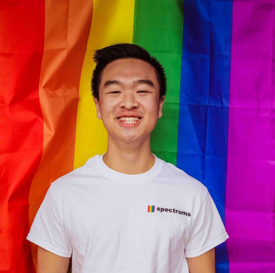 Senior Community Liaison Evan Chan stands in front of the rainbow flag. He identifies as queer. 